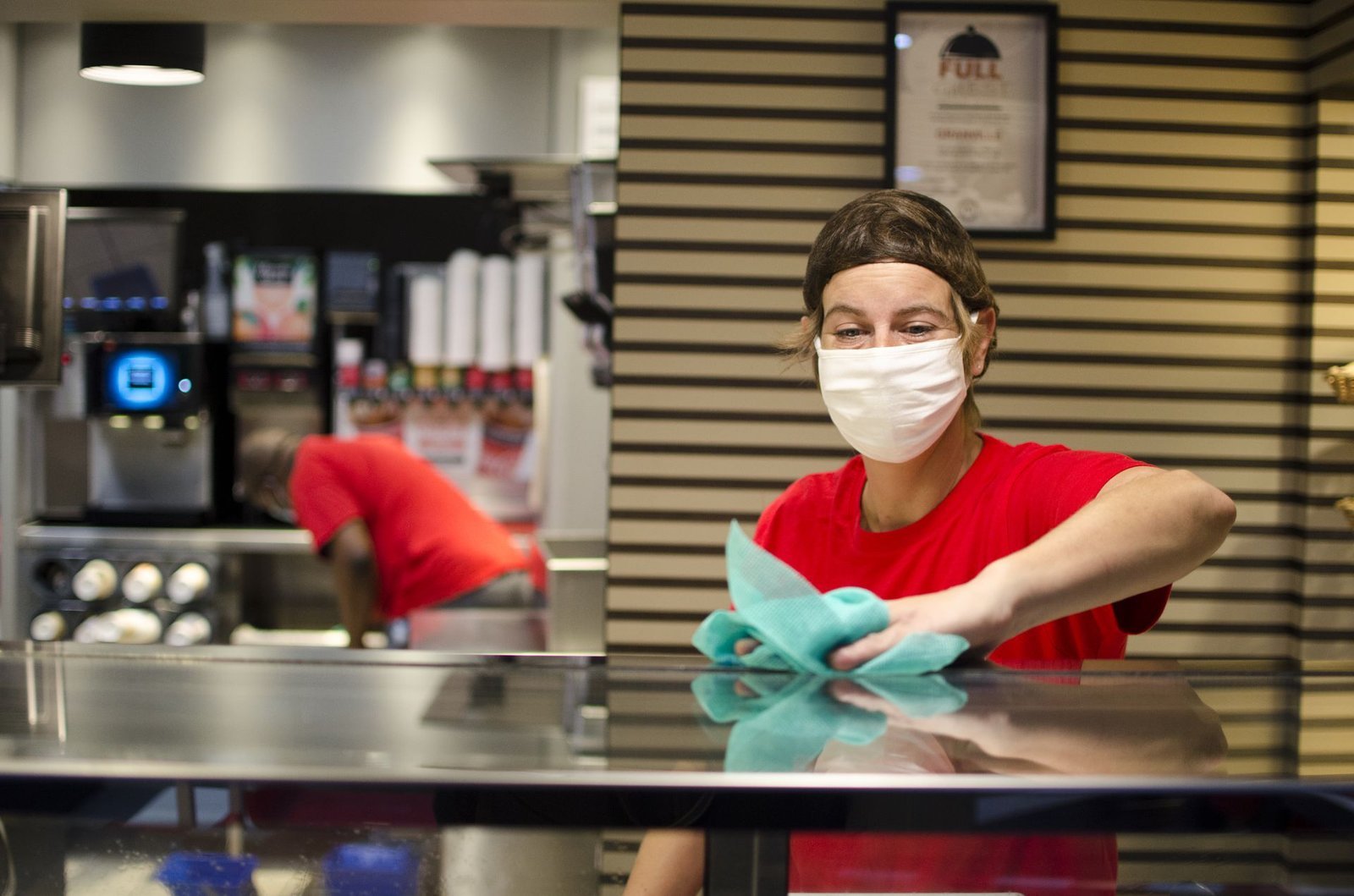 Employée portant un masque nettoyant une surface métallique avec un chiffon dans un espace de restauration.