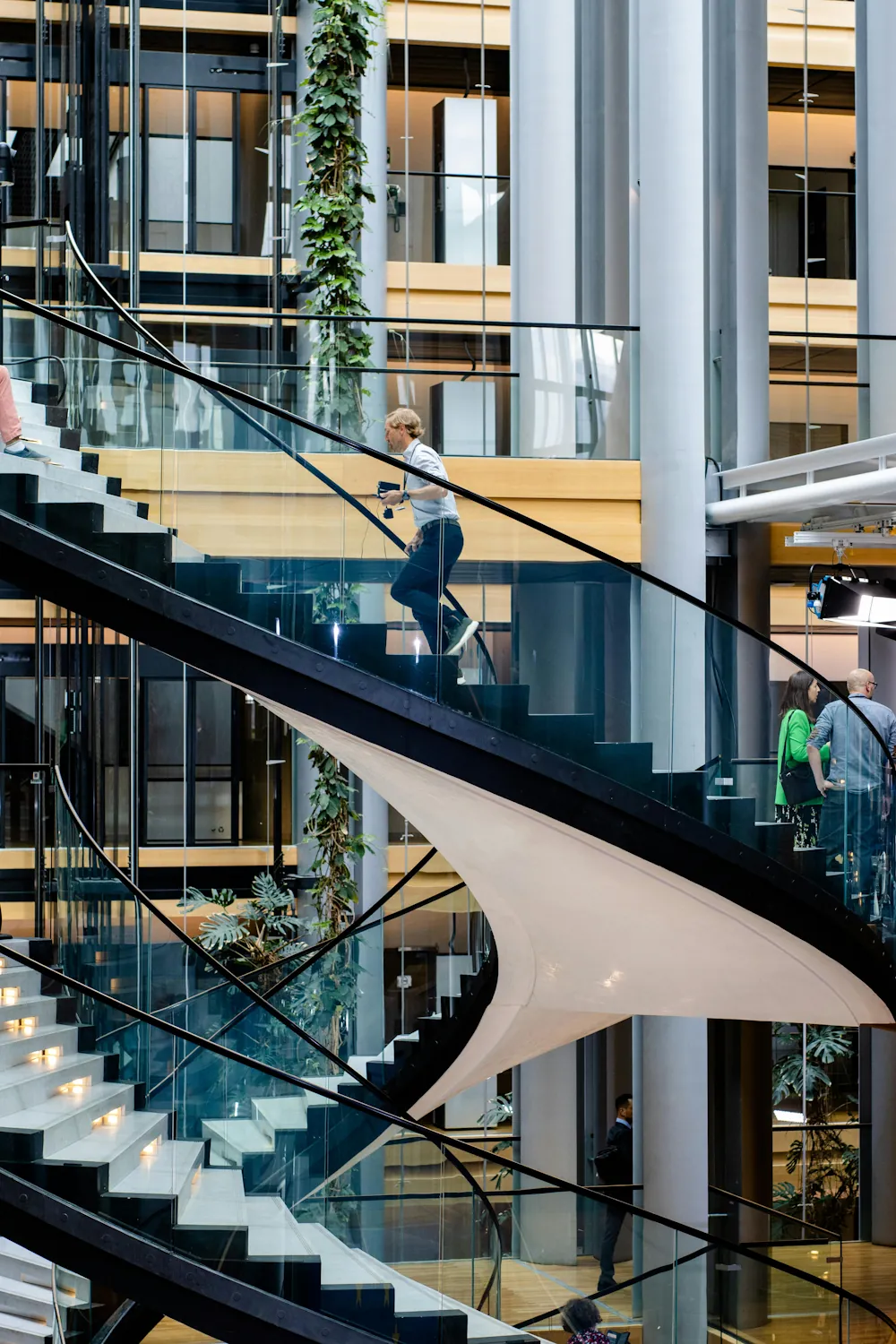 Une personne monte des escaliers dans un bâtiment moderne avec cage d’escalier en verre et structure métallique.