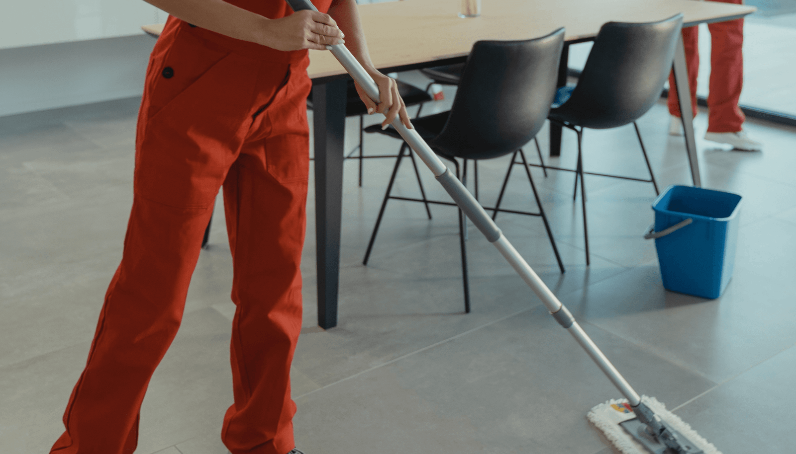 Personne en tenue de travail rouge utilisant un balai plateau sur un sol intérieur.